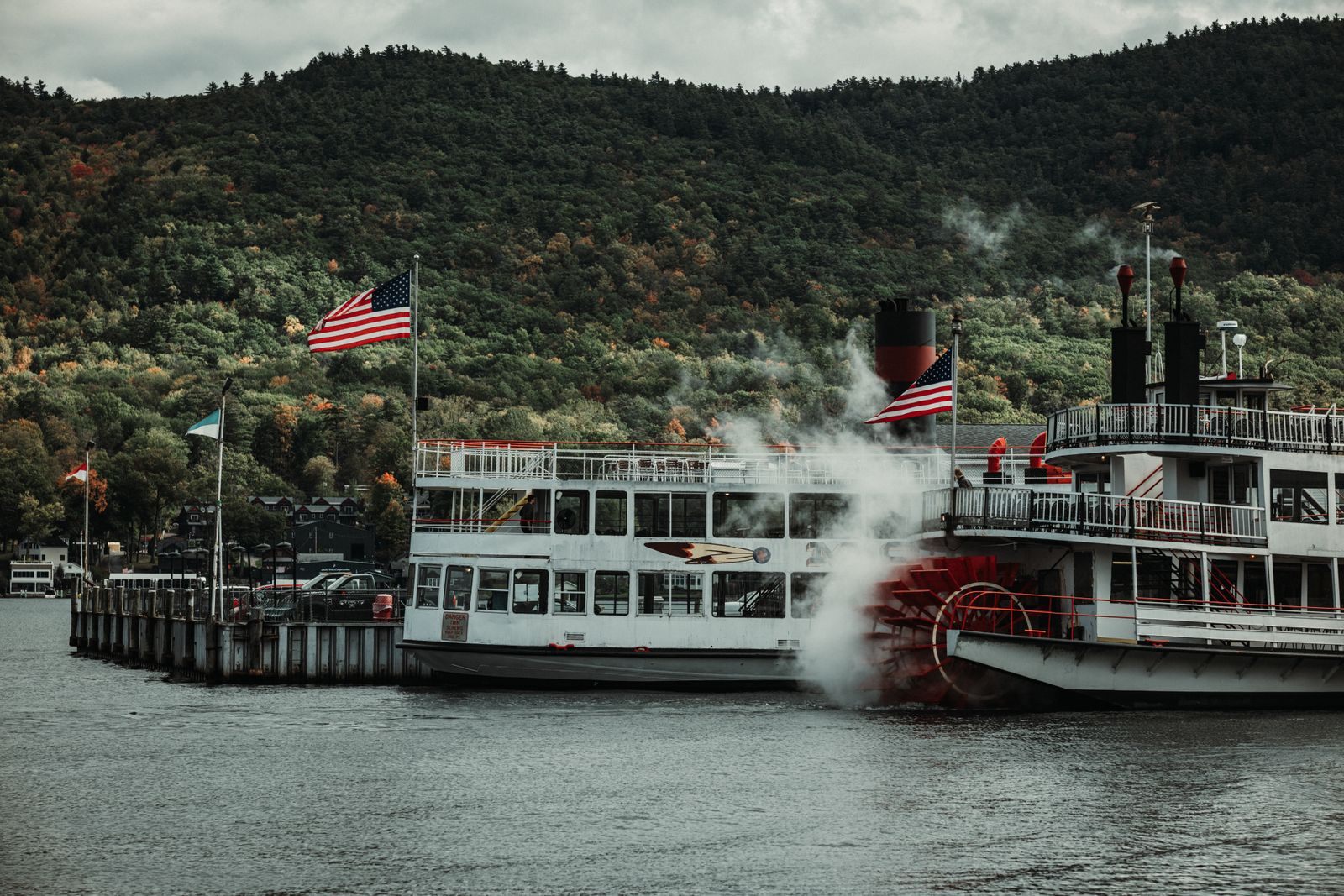 Paddlewheel steamboat with flags flying tied up below wooded autumn hills, steam rising from the stern